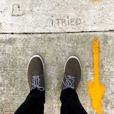 Two shoes from above standing before the words "I tried" carved into the pavement