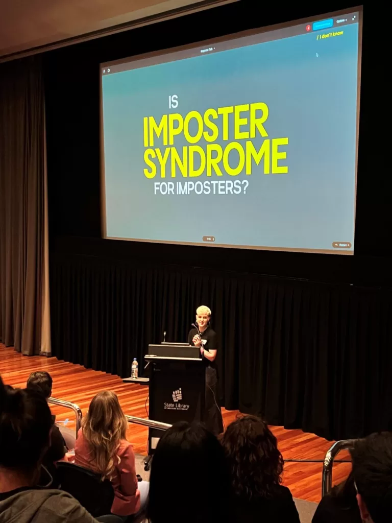 Jess Witt is preparing to start her closing keynote, "Is imposter syndrome for imposters?". She is standing on the stage behind a lecture with the screen behind her.